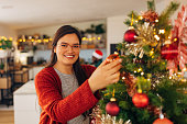 Smiling Asian woman looking directly at the camera while decorating the Christmas tree in the living room of her modern apartment